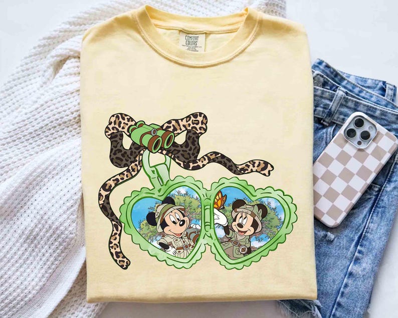 Puede incluir: Una camiseta amarilla con un gr&aacute;fico de dos personajes de dibujos animados en un medall&oacute;n en forma de coraz&oacute;n. El medall&oacute;n est&aacute; rodeado por una cinta de estampado de leopardo marr&oacute;n y negro. Los personajes llevan atuendos de safari y sostienen binoculares. El texto "COMFORT COLORS" es visible en la camiseta.