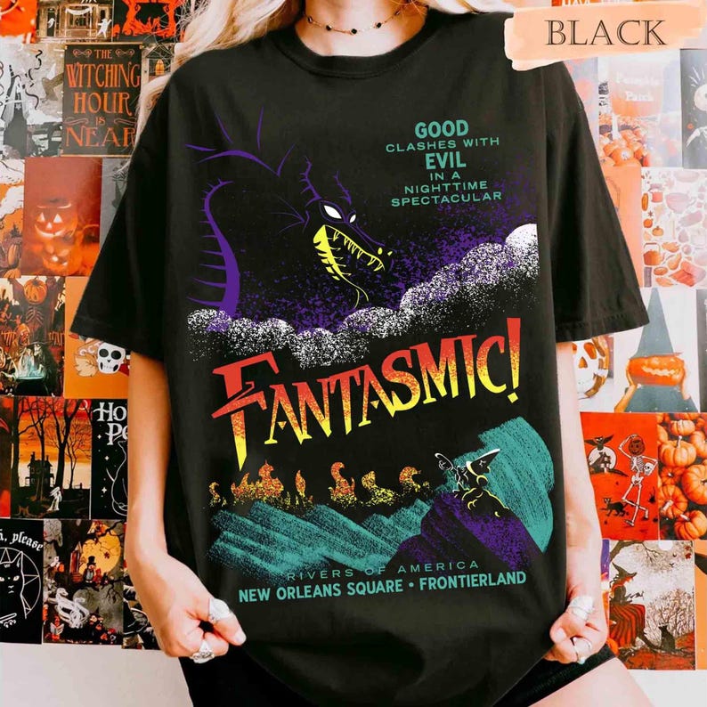 K&ouml;nnte beinhalten: Schwarzes T-Shirt mit einem farbenfrohen Grafikdesign. Das Design zeigt das Wort "FANTASMIC!" in gro&szlig;en, stilisierten Buchstaben, einen Drachen und den Text "GOOD CLASHES WITH EVIL IN A NIGHTTIME SPECTACULAR". Enth&auml;lt auch "RIVERS OF AMERICA" und "NEW ORLEANS SQUARE &bull; FRONTIERLAND".