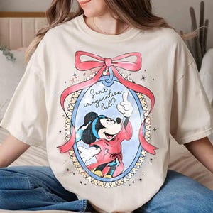 May include: Cream-coloured t-shirt featuring a cartoon image of Mickey Mouse giving a thumbs-up inside an oval frame with a pink bow. The text "Some imagination huh?" is above the image. The shirt is a relaxed fit.