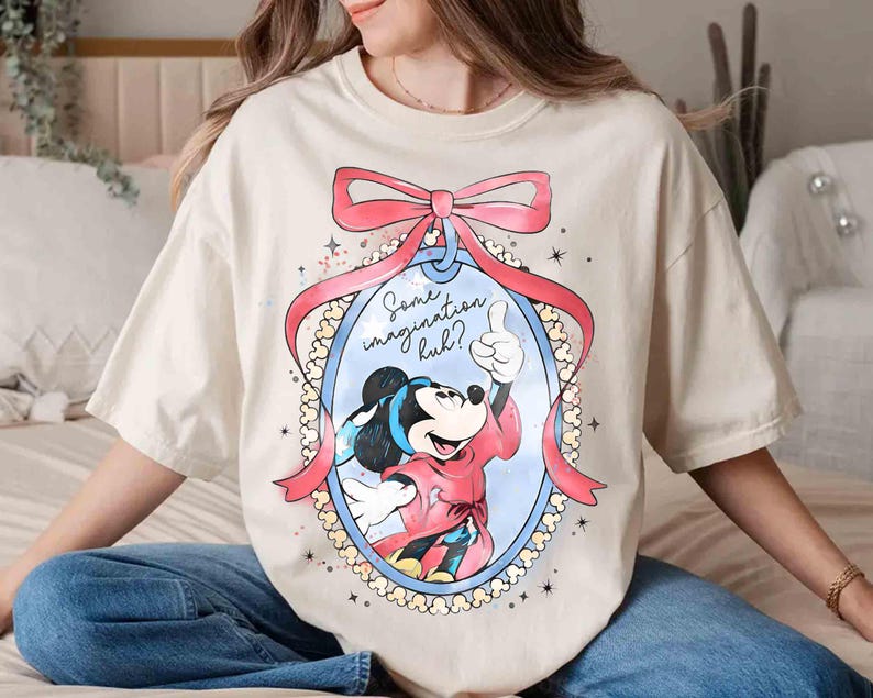 May include: Cream-coloured t-shirt featuring a cartoon image of Mickey Mouse giving a thumbs-up inside an oval frame with a pink bow. The text "Some imagination huh?" is above the image. The shirt is a relaxed fit.