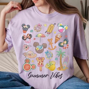 以下が含まれることがあります： 様々な夏のテーマのアイコンがカラフルなグラフィックプリントされたパープルのTシャツ。ミッキーマウスの耳、アイスクリームコーン、ヤシの木、スイカなどがあります。Tシャツの下部に「Summer Vibes」というテキストが黒でプリントされています。