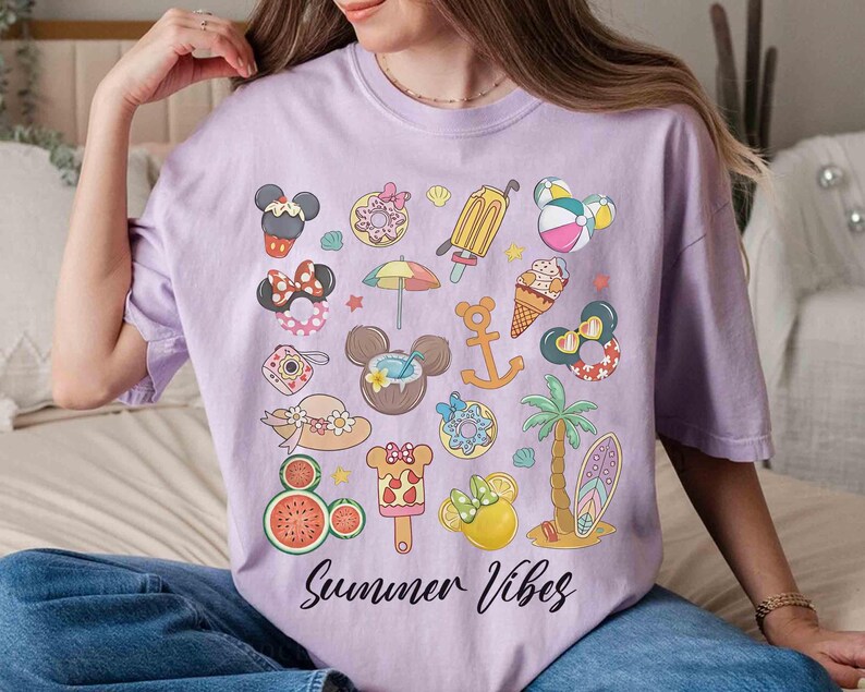 以下が含まれることがあります： 様々な夏のテーマのアイコンがカラフルなグラフィックプリントされたパープルのTシャツ。ミッキーマウスの耳、アイスクリームコーン、ヤシの木、スイカなどがあります。Tシャツの下部に「Summer Vibes」というテキストが黒でプリントされています。