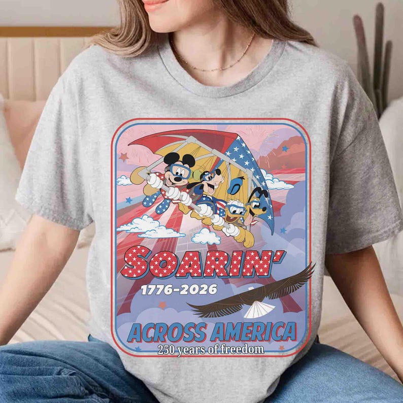 May include: A gray t-shirt featuring a graphic of Mickey Mouse, Goofy, and Donald Duck soaring in a hang glider. The design includes the text "SOARIN' 1776-2026 ACROSS AMERICA 250 years of freedom" with an eagle illustration.