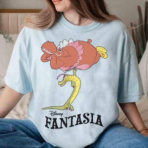 May include: Light blue t-shirt featuring a cartoon illustration of a hippo in a pink tutu and a yellow and green alligator. The word "Disney FANTASIA" is printed in black below the illustration.