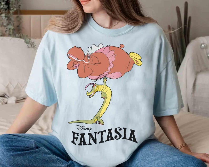 May include: Light blue t-shirt featuring a cartoon illustration of a hippo in a pink tutu and a yellow and green alligator. The word "Disney FANTASIA" is printed in black below the illustration.