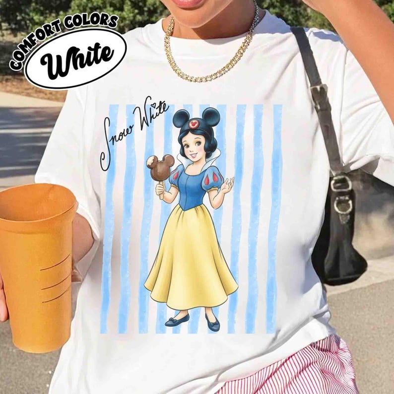 May include: A white t-shirt featuring a graphic of Snow White wearing mouse ears and holding a chocolate treat. The design includes vertical light blue stripes and the text "Snow White" in black script. The shirt has a crew neckline.
