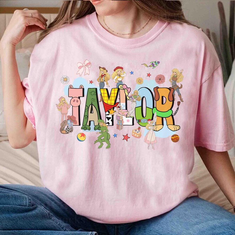 May include: A light pink short-sleeve t-shirt featuring the name TAYLOR in large, colorful, patterned block letters. The design is surrounded by various cartoon characters, small stars, flowers, and shapes in a bright, multi-colored graphic print.