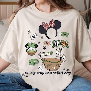 K&ouml;nnte beinhalten: Cremefarbenes T-Shirt mit farbenfrohem Grafikdesign. Das Design umfasst ein Minnie Mouse Ohren-Stirnband, eine Bauchtasche, einen Cupcake und andere thematische Elemente. Der Text "on my way to a safari day" ist auf dem Shirt aufgedruckt.