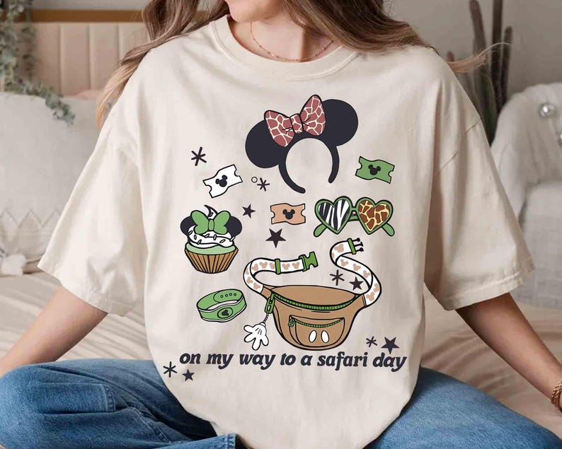 K&ouml;nnte beinhalten: Cremefarbenes T-Shirt mit farbenfrohem Grafikdesign. Das Design umfasst ein Minnie Mouse Ohren-Stirnband, eine Bauchtasche, einen Cupcake und andere thematische Elemente. Der Text "on my way to a safari day" ist auf dem Shirt aufgedruckt.