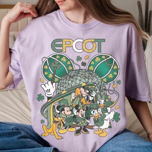 May include: A light purple t-shirt featuring the word "EPCOT" in green and gold lettering. The graphic includes Mickey Mouse, Minnie Mouse, Donald Duck, and Pluto in St. Patrick's Day attire, with shamrocks and a stylized Spaceship Earth.