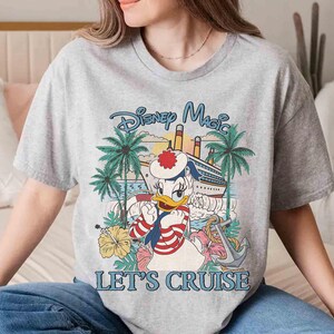 May include: A gray t-shirt featuring a Disney Magic design with Donald Duck in a sailor outfit, a cruise ship, palm trees, and the text "LET'S CRUISE". The graphic is in shades of blue, red, yellow, and green.