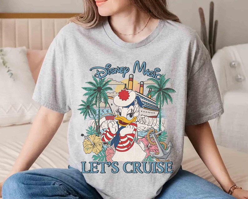 May include: A gray t-shirt featuring a Disney Magic design with Donald Duck in a sailor outfit, a cruise ship, palm trees, and the text "LET'S CRUISE". The graphic is in shades of blue, red, yellow, and green.