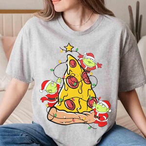 Op de afbeelding: Grijs t-shirt met een pizzapunt versierd als een kerstboom. De pizza heeft pepperoni en is versierd met lichtslingers en een ster. Drie groene aliens met kerstmutsen versieren de pizza.