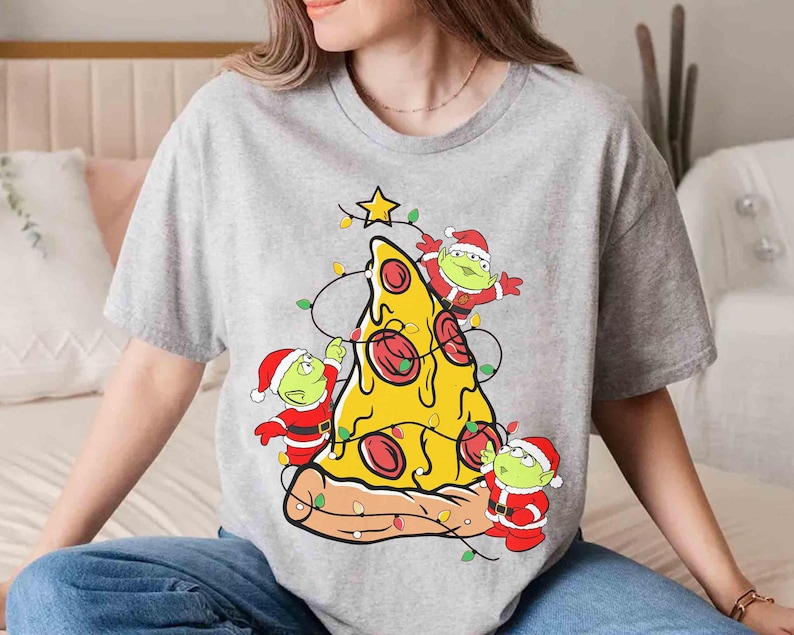 Op de afbeelding: Grijs t-shirt met een pizzapunt versierd als een kerstboom. De pizza heeft pepperoni en is versierd met lichtslingers en een ster. Drie groene aliens met kerstmutsen versieren de pizza.