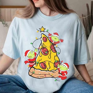 Op de afbeelding: Lichtblauw T-shirt met een pizzapunt versierd als kerstboom. De pizza heeft pepperoni en is versierd met lichtsnoeren en een ster. Drie groene aliens in kerstmanpakken versieren de pizza.