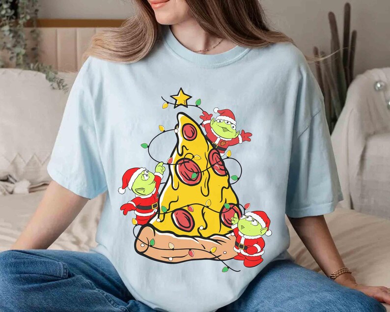 Op de afbeelding: Lichtblauw T-shirt met een pizzapunt versierd als kerstboom. De pizza heeft pepperoni en is versierd met lichtsnoeren en een ster. Drie groene aliens in kerstmanpakken versieren de pizza.