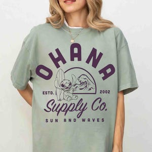 May include: Sage green t-shirt with a purple graphic featuring the word "OHANA" above a cartoon character with a surfboard and a wave. The text "Supply Co. SUN AND WAVES" is below the graphic. The shirt also has the text "ESTD. 2002".