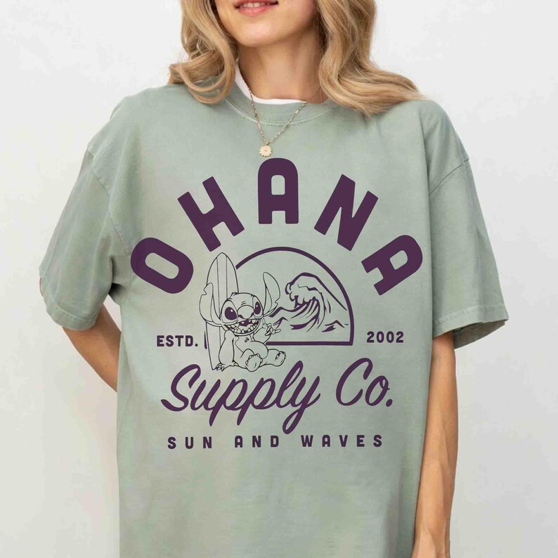 May include: Sage green t-shirt with a purple graphic featuring the word "OHANA" above a cartoon character with a surfboard and a wave. The text "Supply Co. SUN AND WAVES" is below the graphic. The shirt also has the text "ESTD. 2002".