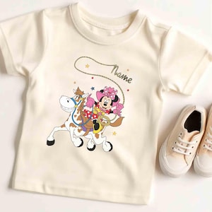 May include: Cream-colored t-shirt featuring a cartoon Minnie Mouse riding a horse with a lasso. The design includes stars and the word "Name". A pair of cream-colored sneakers are next to the shirt.