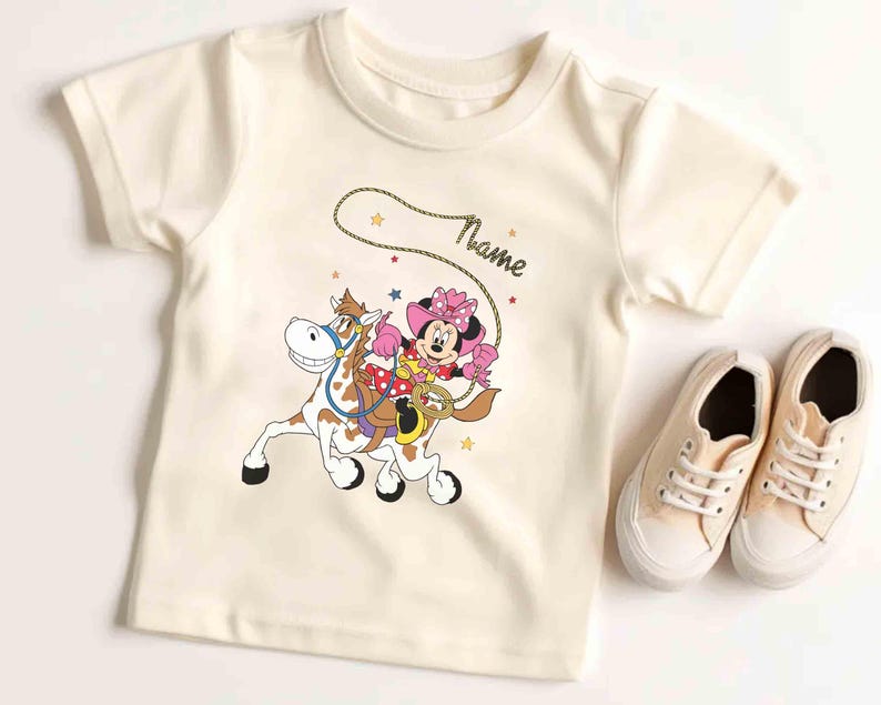 May include: Cream-colored t-shirt featuring a cartoon Minnie Mouse riding a horse with a lasso. The design includes stars and the word "Name". A pair of cream-colored sneakers are next to the shirt.
