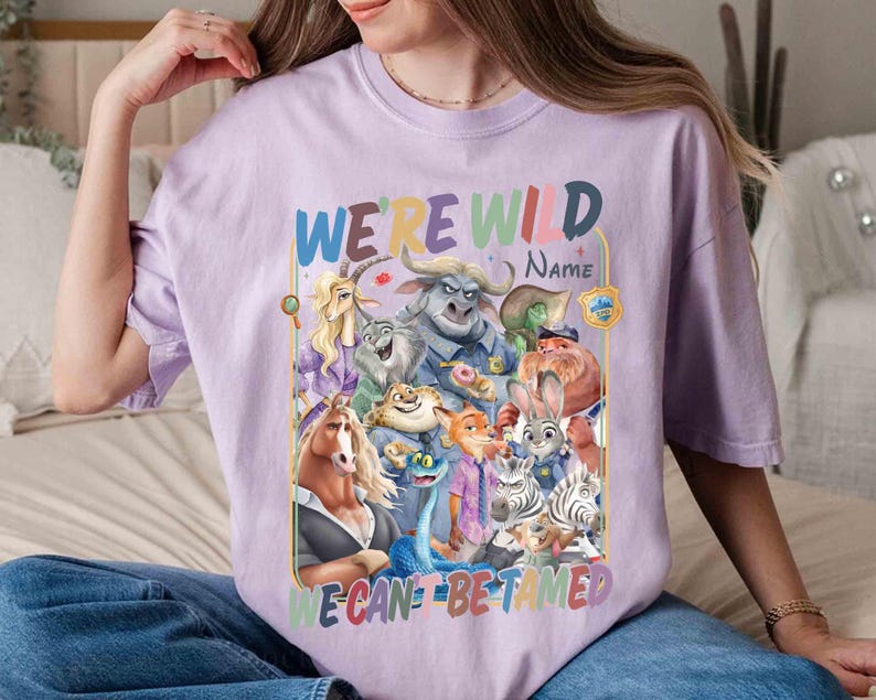 May include: A lilac-colored t-shirt featuring a colorful graphic of animated characters. The text "WE'RE WILD" and "WE CAN'T BE TAMED" is displayed above and below the characters. The word "NAME" is also present.