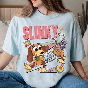 May include: Light blue t-shirt featuring a graphic of Slinky Dog from Toy Story. The design includes the text "SLINKY DOG" in pink and "TOY STORY" with "SINCE 1995". The cartoon dog is brown, tan, and yellow.