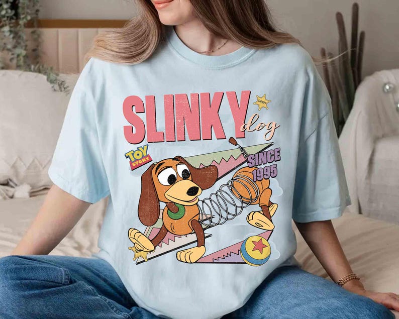 May include: Light blue t-shirt featuring a graphic of Slinky Dog from Toy Story. The design includes the text "SLINKY DOG" in pink and "TOY STORY" with "SINCE 1995". The cartoon dog is brown, tan, and yellow.