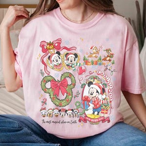 May include: Pink t-shirt featuring a festive Christmas design with Mickey Mouse, Minnie Mouse, Chip and Dale, and other Disney characters. The shirt includes the text "Very Merry Christmas" and "The most magical place on Earth."