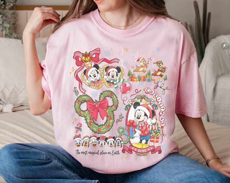 May include: Pink t-shirt featuring a festive Christmas design with Mickey Mouse, Minnie Mouse, Chip and Dale, and other Disney characters. The shirt includes the text "Very Merry Christmas" and "The most magical place on Earth."