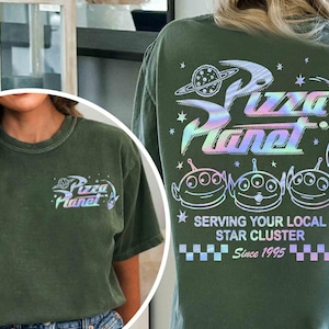 May include: Olive green t-shirt featuring the Pizza Planet logo with a holographic design on the front and back. The back of the shirt displays the Pizza Planet logo, three alien characters, and the text "Serving Your Local Star Cluster Since 1995."