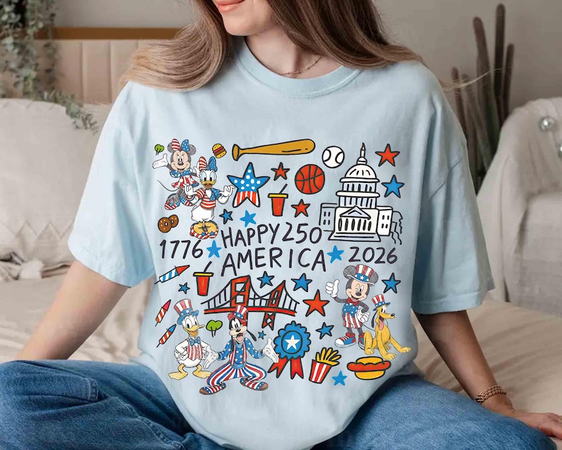 Puede incluir: Camiseta azul claro con un gr&aacute;fico colorido que celebra el 250 aniversario de Am&eacute;rica. El dise&ntilde;o incluye personajes de dibujos animados, el Capitolio, un bate de b&eacute;isbol, estrellas y el texto "1776 HAPPY 250 AMERICA 2026".