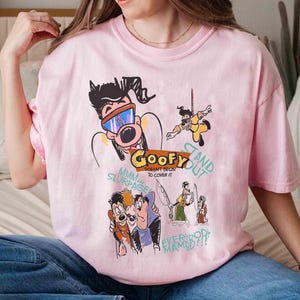 May include: Pink t-shirt featuring a cartoon illustration of Goofy and other characters. The text on the shirt reads "Goofy Doesn't Begin To Cover It" and "Stand Out".