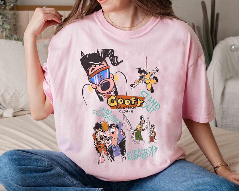 May include: Pink t-shirt featuring a cartoon illustration of Goofy and other characters. The text on the shirt reads "Goofy Doesn't Begin To Cover It" and "Stand Out".