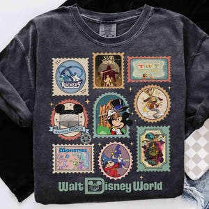 Puede incluir: Camiseta gris oscuro con dise&ntilde;os de sellos postales tem&aacute;ticos de Disney. Los sellos incluyen im&aacute;genes de Rockers, Star Wars, Toy Story, Mickey Mouse, Monsters y m&aacute;s. Las palabras "Walt Disney World" est&aacute;n impresas en la parte inferior.