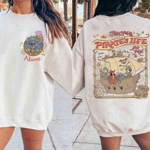K&ouml;nnte beinhalten: Cremefarbenes Sweatshirt mit einer Cartoon-Grafik eines blauen Alien-Charakters mit Piratenhut und dem Wort "Name" darunter. Auf der R&uuml;ckseite des Sweatshirts ist ein Piratenschiff mit Figuren und dem Text "Yo Ho, Pirate's Life for Me!" abgebildet.
