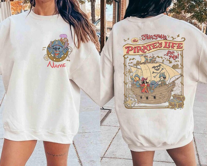 K&ouml;nnte beinhalten: Cremefarbenes Sweatshirt mit einer Cartoon-Grafik eines blauen Alien-Charakters mit Piratenhut und dem Wort "Name" darunter. Auf der R&uuml;ckseite des Sweatshirts ist ein Piratenschiff mit Figuren und dem Text "Yo Ho, Pirate's Life for Me!" abgebildet.