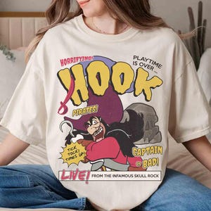 May include: Cream-colored t-shirt featuring a graphic of Captain Hook from Peter Pan. The design includes the word "HOOK" in yellow, a purple pirate hat, and the phrases "HORRIFYING! PIRATES!" and "CAPTAIN OF BAD!"