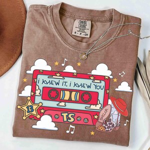 May include: A brown t-shirt featuring a graphic of a red cassette tape with the text "I KNEW IT, I KNEW YOU" and "TS". The design includes white clouds, yellow stars, musical notes, a sheriff star with the number 13, and a cowboy boot with a hat.