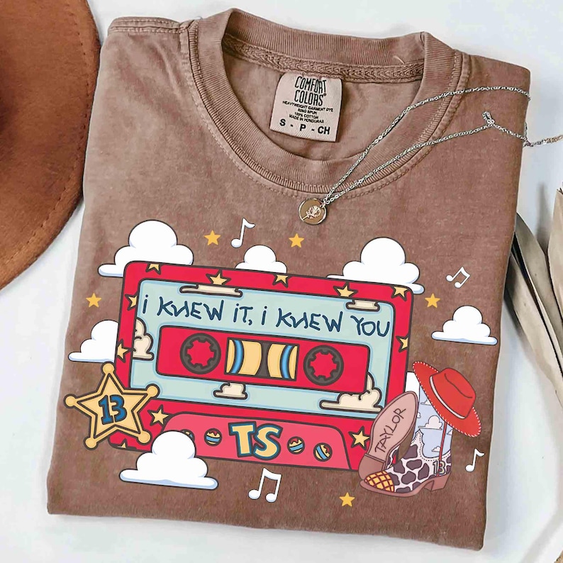 May include: A brown t-shirt featuring a graphic of a red cassette tape with the text "I KNEW IT, I KNEW YOU" and "TS". The design includes white clouds, yellow stars, musical notes, a sheriff star with the number 13, and a cowboy boot with a hat.