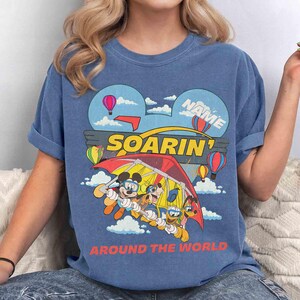 May include: Blue t-shirt with a colorful graphic featuring Mickey Mouse, Goofy, and Donald Duck in a hang glider. The design includes hot air balloons, clouds, and the words "SOARIN'" and "AROUND THE WORLD."