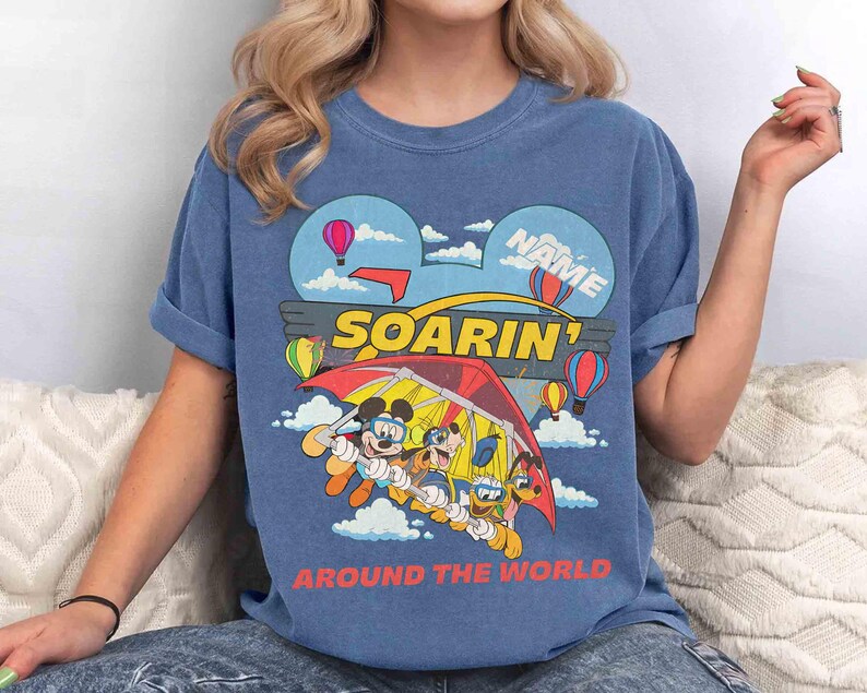 May include: Blue t-shirt with a colorful graphic featuring Mickey Mouse, Goofy, and Donald Duck in a hang glider. The design includes hot air balloons, clouds, and the words "SOARIN'" and "AROUND THE WORLD."