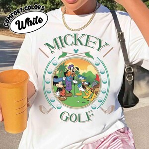 May include: White t-shirt with a circular graphic featuring Mickey Mouse, Goofy, Donald Duck, and Pluto playing golf. The word "MICKEY" is arched above the scene, with "GOLF" below. The shirt also has the words "COMFORT COLORS" and "White" in a white oval.