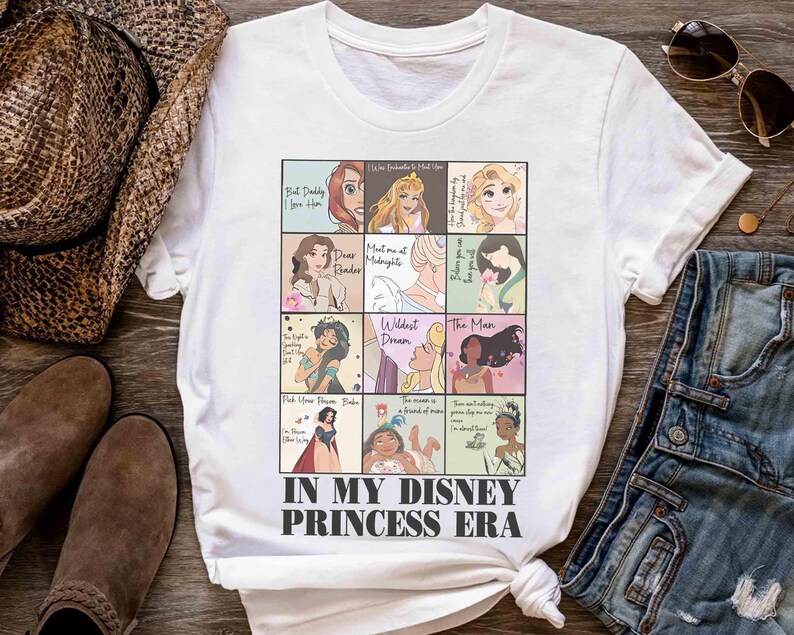May include: White t-shirt featuring a grid of Disney princess illustrations with text. The shirt includes the phrase "IN MY DISNEY PRINCESS ERA" in black font. The shirt is displayed on a wooden surface with accessories.