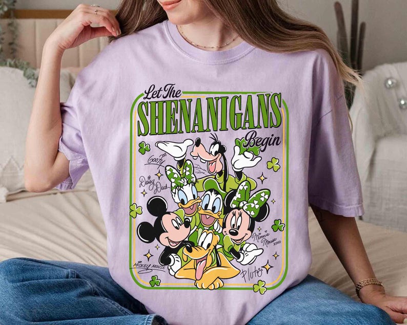 May include: A lilac-colored t-shirt featuring a graphic of Disney characters, including Mickey Mouse, Minnie Mouse, Donald Duck, Daisy Duck, Goofy, and Pluto. The design includes the text "Let The Shenanigans Begin" in a green frame with shamrocks.