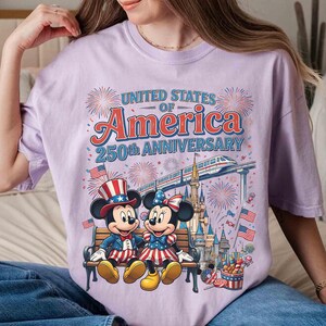 Może przedstawiać: Jasnofioletowa koszulka z Myszką Miki i Minnie w patriotycznych strojach, siedzących na ławce. Na koszulce widnieje napis "UNITED STATES OF AMERICA 250TH ANNIVERSARY" z fajerwerkami i pociągiem.