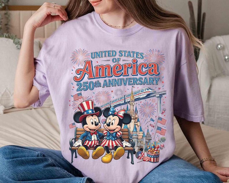Może przedstawiać: Jasnofioletowa koszulka z Myszką Miki i Minnie w patriotycznych strojach, siedzących na ławce. Na koszulce widnieje napis "UNITED STATES OF AMERICA 250TH ANNIVERSARY" z fajerwerkami i pociągiem.