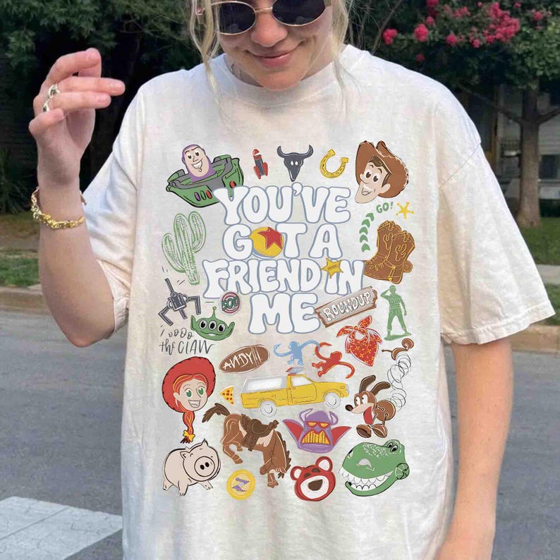 May include: Cream-colored t-shirt featuring a colorful graphic design with characters and text from the movie "Toy Story." The text reads "You've Got a Friend in Me." The design includes images of Buzz Lightyear, Woody, and other characters.