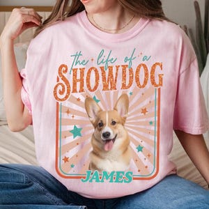 May include: Pink t-shirt with a graphic of a smiling Corgi dog and the text "the life of a SHOWDOG JAMES" in a retro design. The design includes stars and a sunburst pattern, creating a vintage aesthetic.
