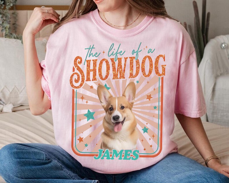 May include: Pink t-shirt with a graphic of a smiling Corgi dog and the text "the life of a SHOWDOG JAMES" in a retro design. The design includes stars and a sunburst pattern, creating a vintage aesthetic.