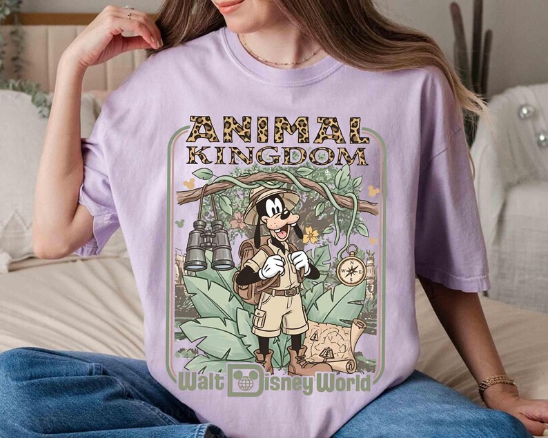 Puede incluir: Una camiseta lila con un dise&ntilde;o de Goofy con ropa de safari, el texto "ANIMAL KINGDOM" y "Walt Disney World". El dise&ntilde;o incluye binoculares, una br&uacute;jula y un mapa, sobre un fondo de jungla.